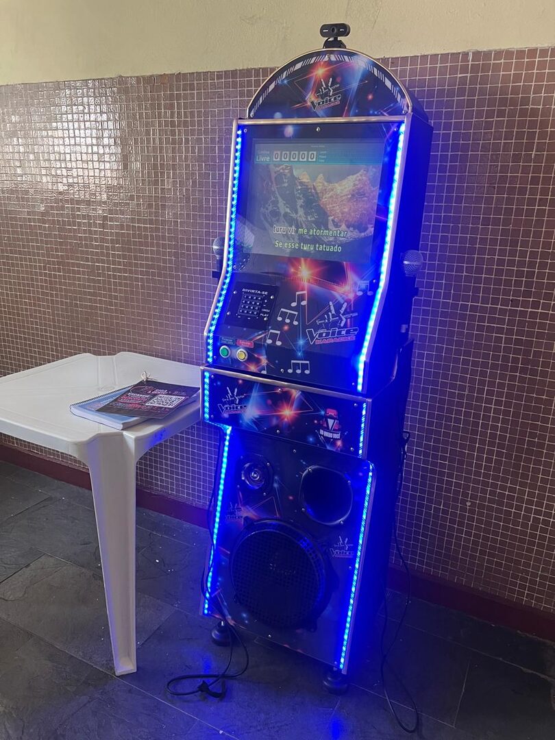 Máquina de karaokê DanSan com LED azul ligada, tocando uma música na tela, ao lado de uma mesa com o catálogo de músicas